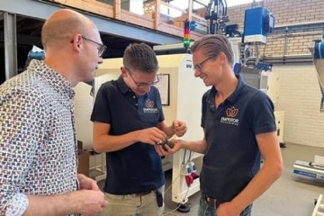 Trzech mężczyzn w hali produkcyjnej omawia detal z tworzywa sztucznego wyprodukowany na wtryskarce znajdującej się za nimi.