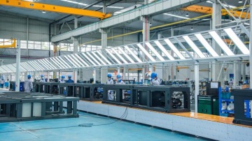 Wnętrze hali przemysłowej z linią montażową, stanowiskami roboczymi oraz pracownikami w odzieży ochronnej.