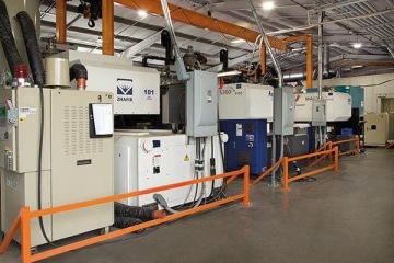 Hala przemysłowa z rzędem wtryskarek Zhafir, zabezpieczona pomarańczowymi barierami, z suwnicą i przewodami wentylacyjnymi.