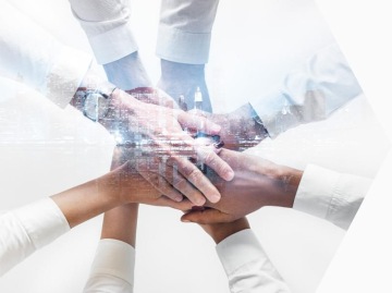 Zbliżenie na dłonie kilku osób ułożone jedna na drugiej; osoby ubrane w koszule, na obrazie widoczna subtelna nakładka miejskiej panoramy.