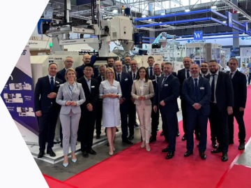 Grupa przedstawicieli firm Mapro i Haitian w strojach biznesowych stojąca na stoisku targowym w hali wystawowej, w tle maszyny przemysłowe.