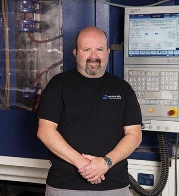 Mężczyzna z brodą w czarnej koszulce stojący z założonymi rękami przed niebieską wtryskarką z panelem sterowania.