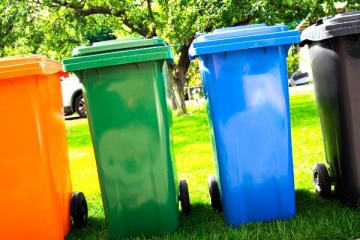 Cztery kolorowe pojemniki na śmieci z otwartymi klapami ustawione w rzędzie na zielonej trawie z drzewami oraz samochodem w tle.