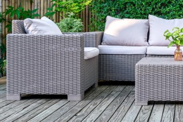 Zestaw ogrodowych mebli z technorattanu z poduszkami składający się z sofy i stolika ustawiony na tarasie z roślinami i bambusowym płotem w tle.