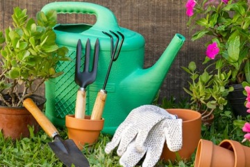 Akcesoria ogrodnicze, w tym zielona konewka, rękawice, łopatka i grabki, ułożone wśród roślin i doniczek w ogrodzie.