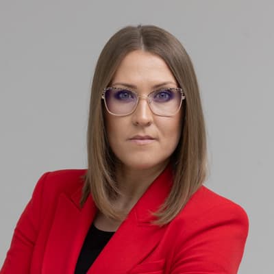 Portret kobiety w czerwonej marynarce i okularach, patrzącej prosto w obiektyw, na jasnym tle.